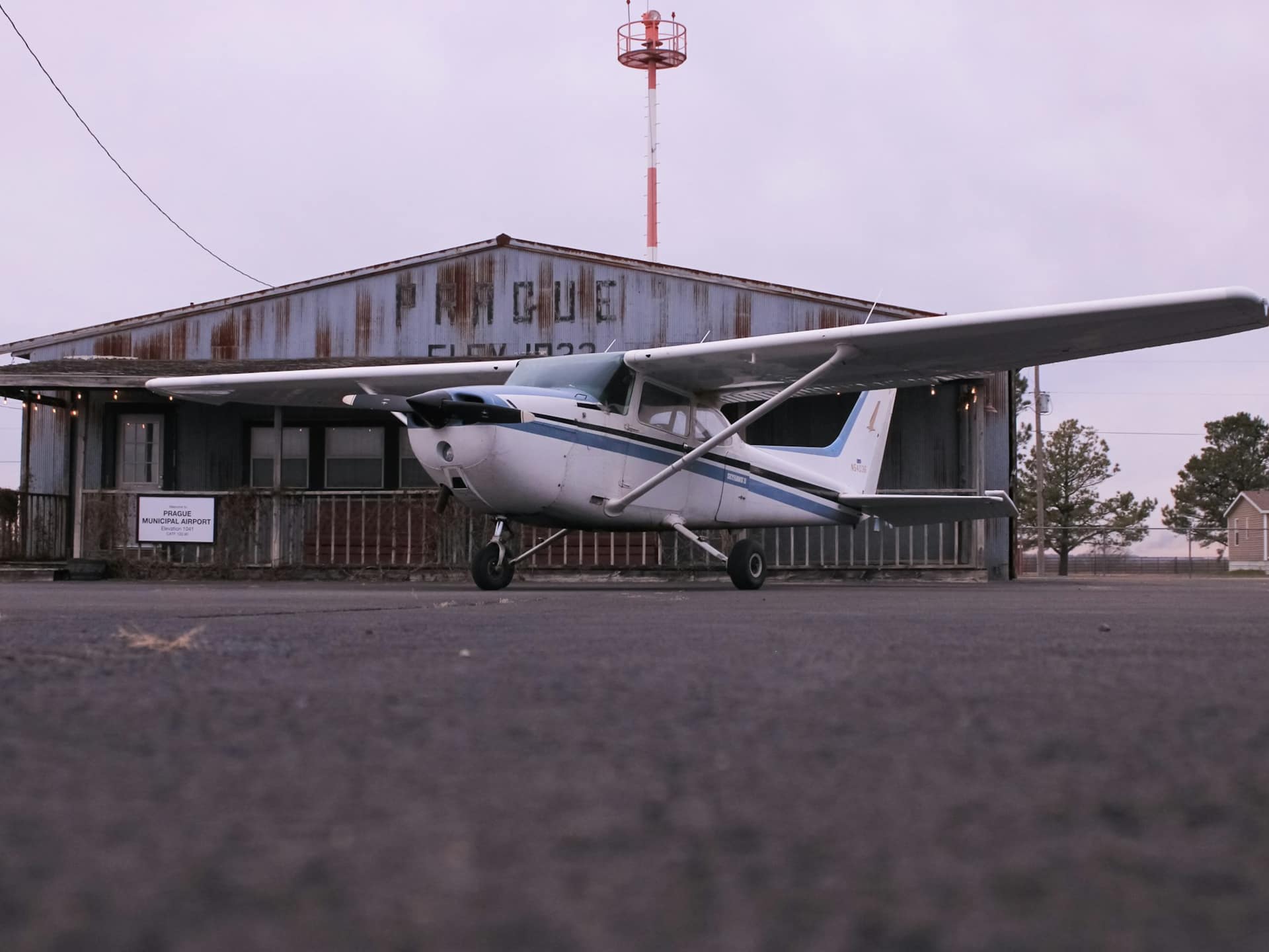 A white Cessna 172 parked on an airfield apron in front of a weathered wooden building