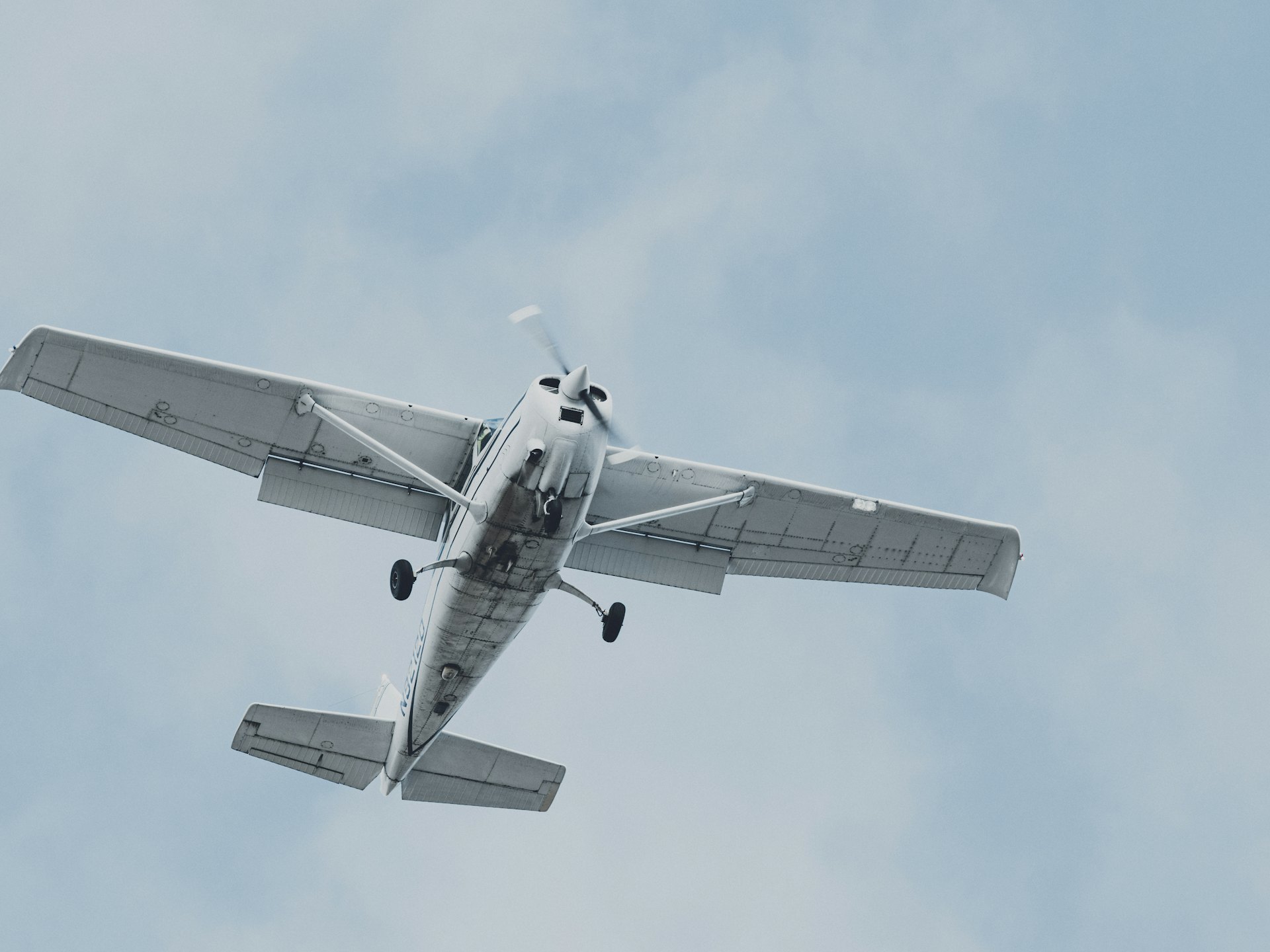 A high-wing Cessna passing overhead, seen from directly below against a pale overcast sky