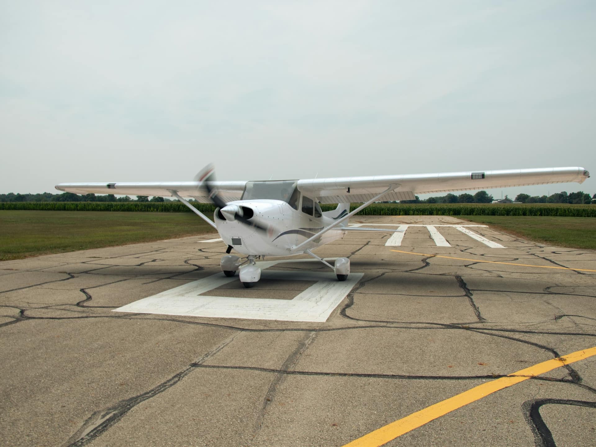 A Cessna 172 with its propeller turning, holding on a runway beside green fields